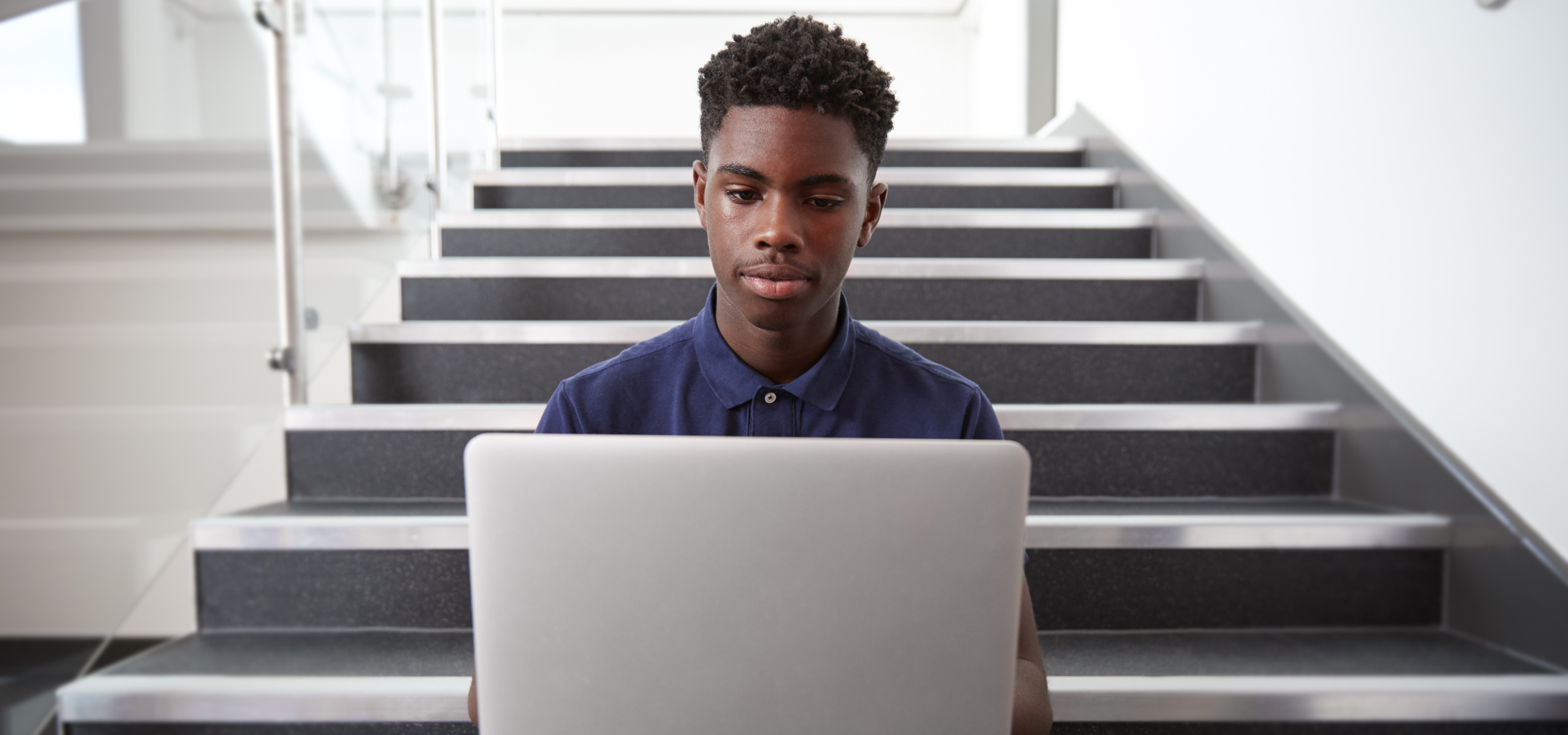 Male high school student sitting on a staircase and using a laptop computer.